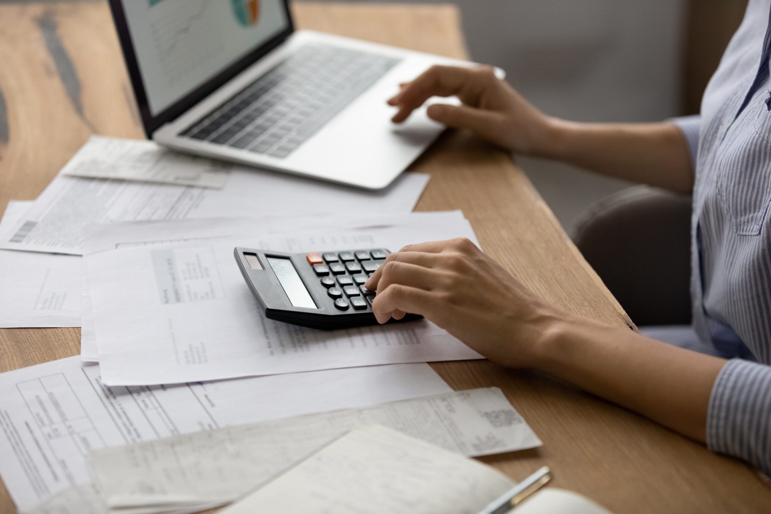 Crop,Close,Up,Of,Caucasian,Woman,Sit,At,Desk,Calculate Person using a calculator and laptop at a desk covered with invoices, receipts, and documents, likely working on finances or budgeting.