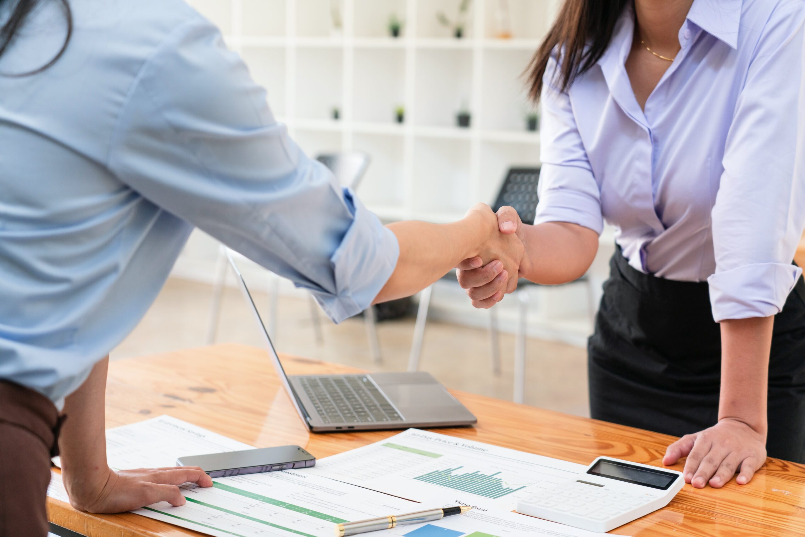 Two,Businesswomen,Shaking,Hands,After,Reading,Paper,And,Discussion,Deal Two people in business attire shake hands over a desk with documents, a laptop, and a calculator, suggesting a professional agreement or meeting.